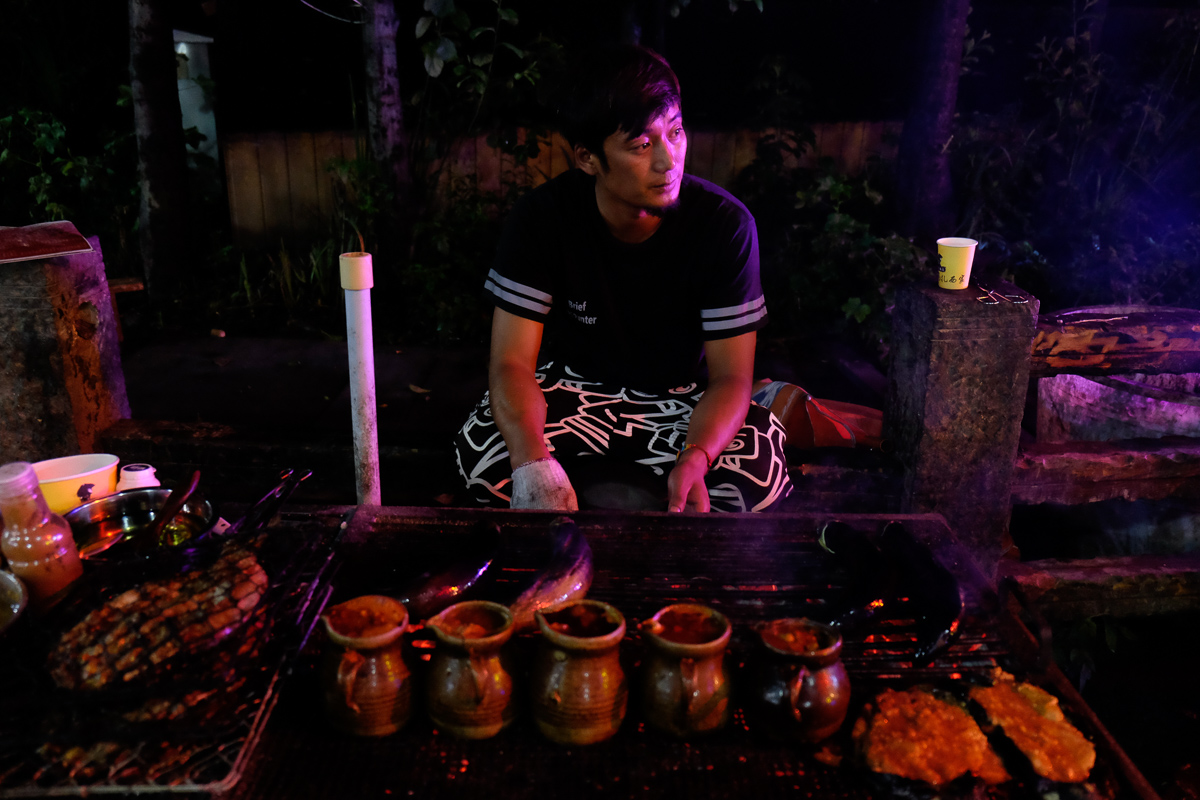 A cook prepares grilled fish and eggplant and little pots of pigs brain at a street-side barbecue restaurant